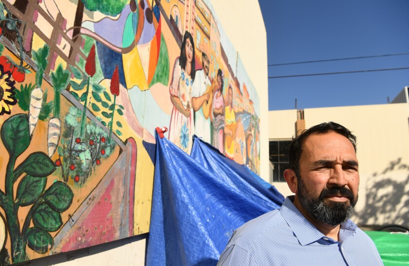A man stands in front of a Mexican cultural mural, with tarps from a homeless camp nearby