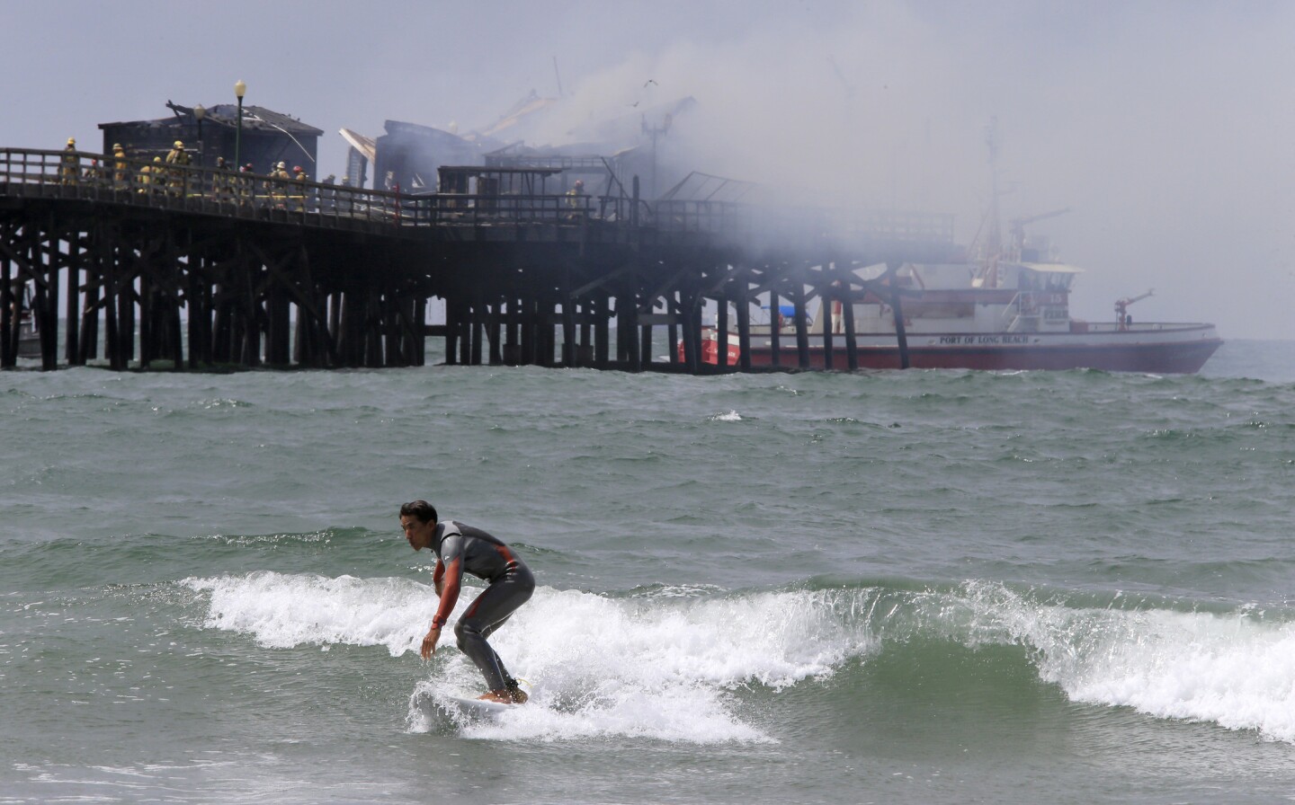 Firefighters mop up a blaze at the end of the Seal Beach Pier Friday morning as a surfer catches a wave.
