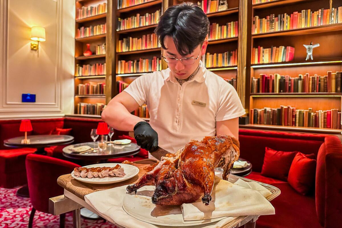 A restaurant server carves Peking duck in a room with red velvet banquettes and book-lined shelves.