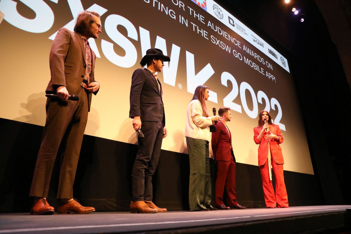 Five men and women stand on a stage in front of a movie screen.