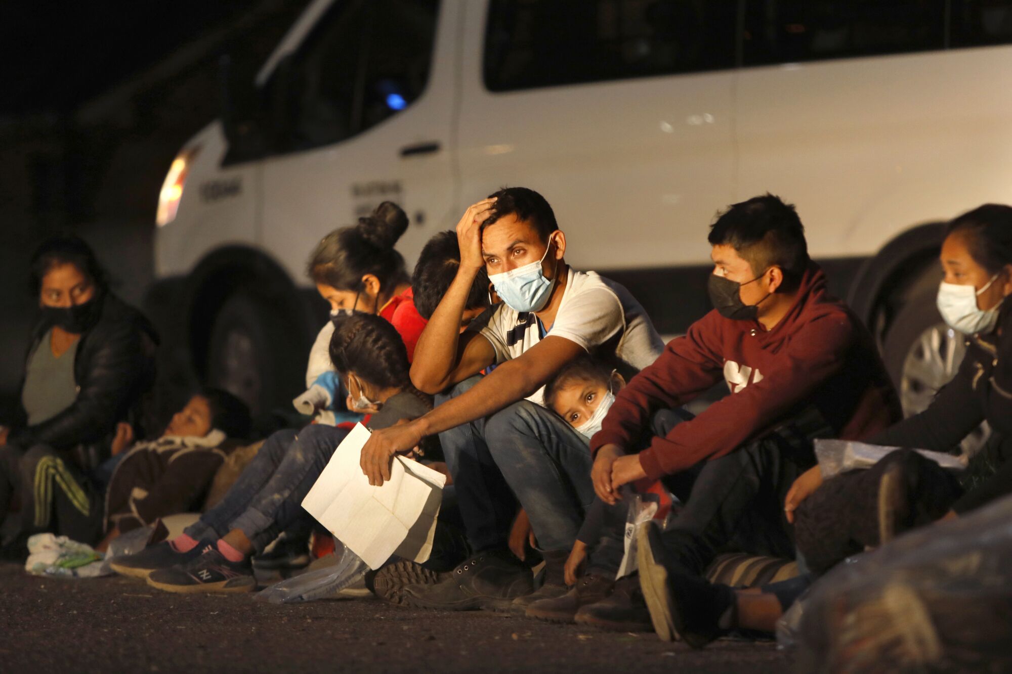 A group of migrant adults and children sit on the ground next to a van at night