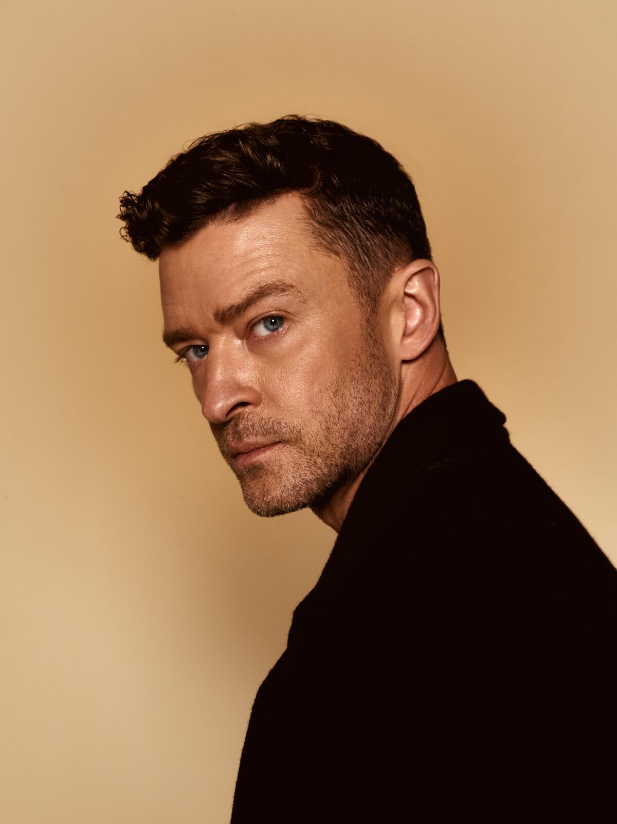 Justin Timberlake in black looks over his left shoulder as he poses against a solid light yellow backdrop