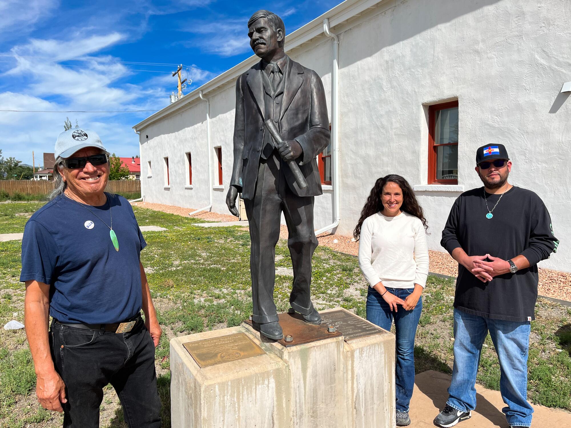 A man, left, and a woman and another man flank a statue of a man near a white building