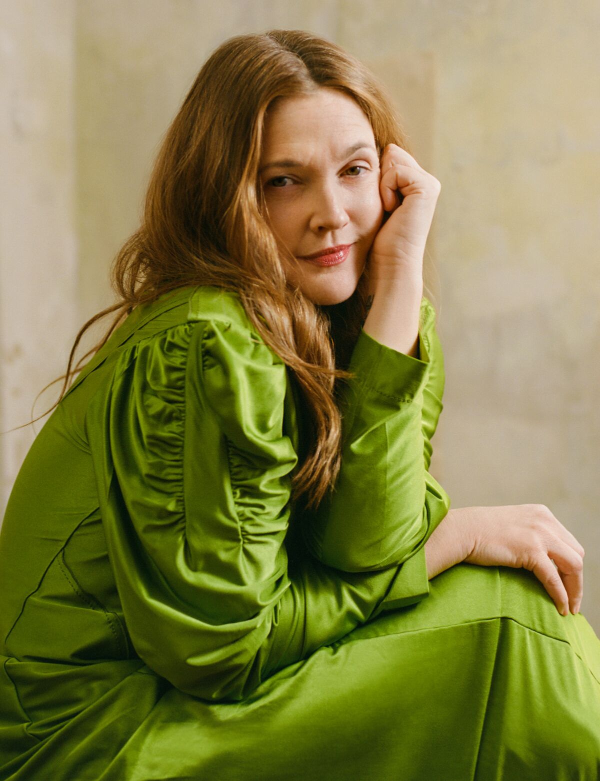 A woman in a shiny green outfit sits sideways with arms crossed and one hand by her left cheek