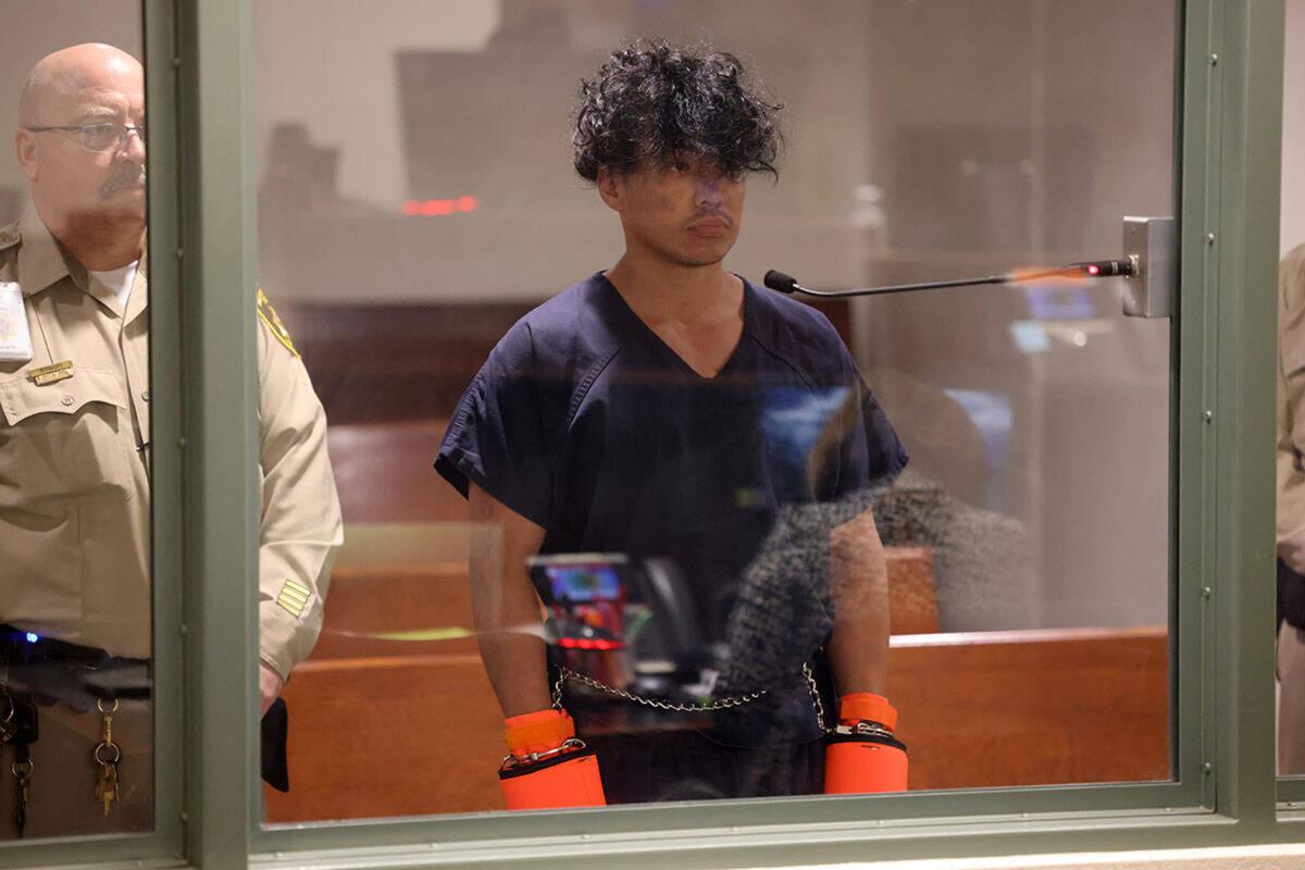 A man in handcuffs and jail mittens stands behind plexiglass in a courtroom