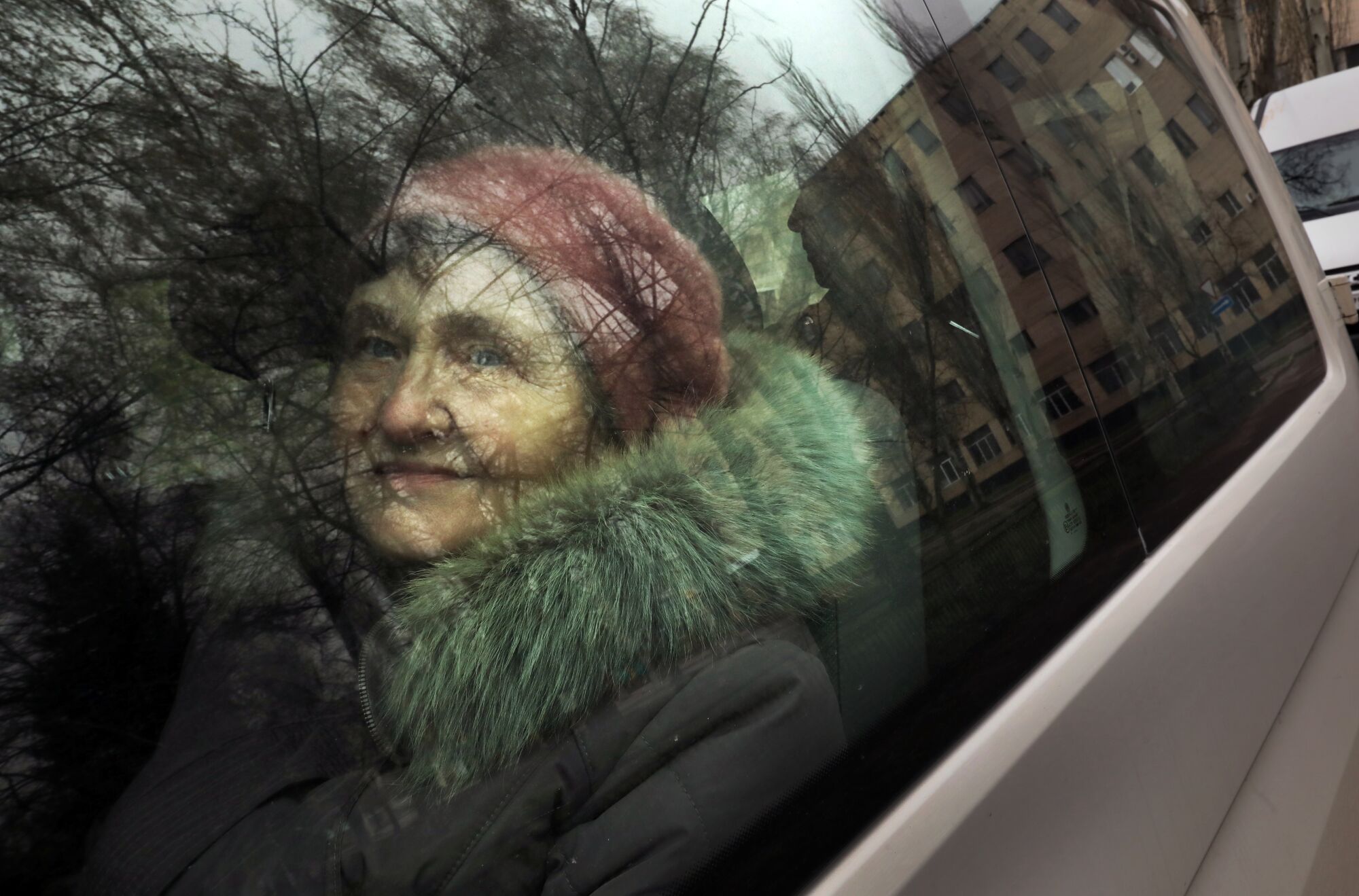 A woman looks out a vehicle window, which also reflects the trees outside