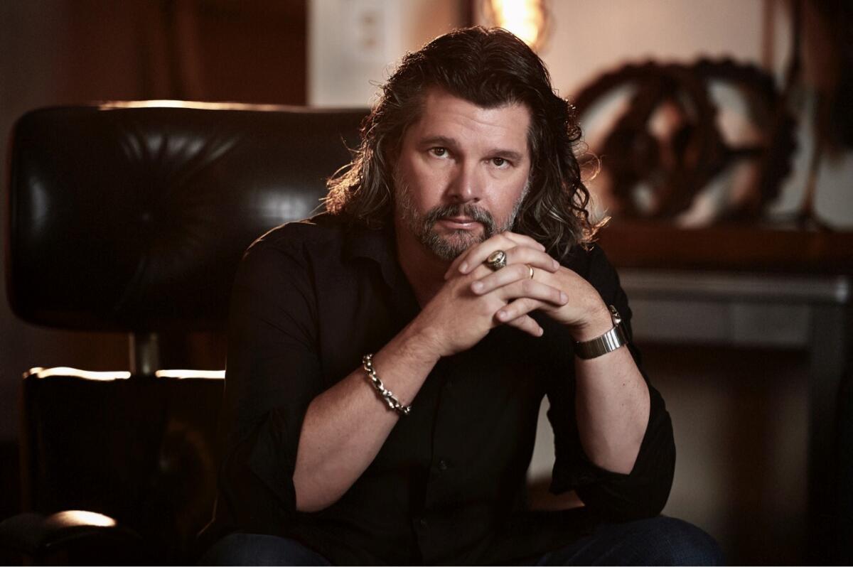 Closeup portrait of a man with collar-length hair and a beard, seated with his elbows on his knees and hands clasped.