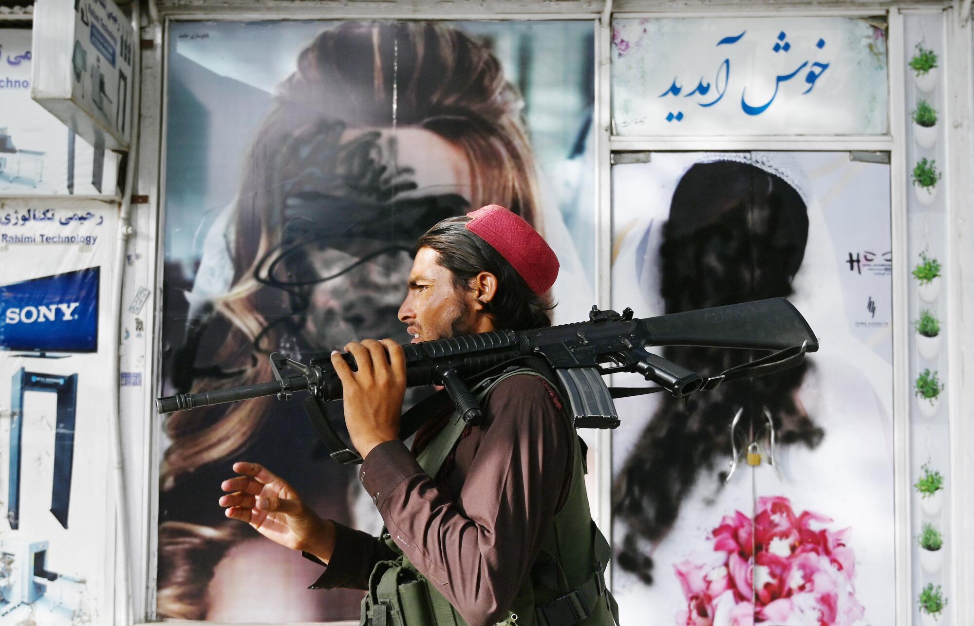 A Taliban fighter holding a gun on his shoulder walks past images of women that have been obscured by paint.