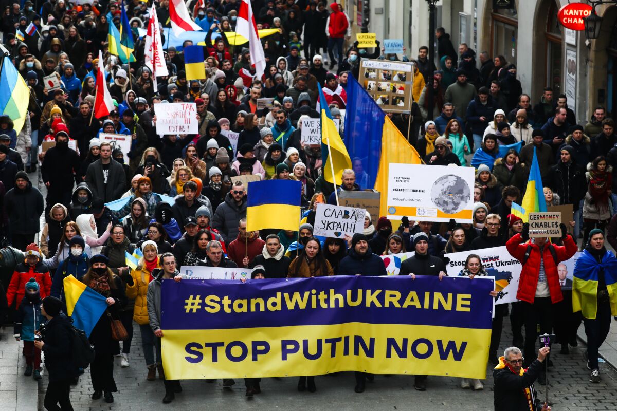 People holding a banner that reads "#StandWithUkraine Stop Putin Now" is joined by others holding colorful flags