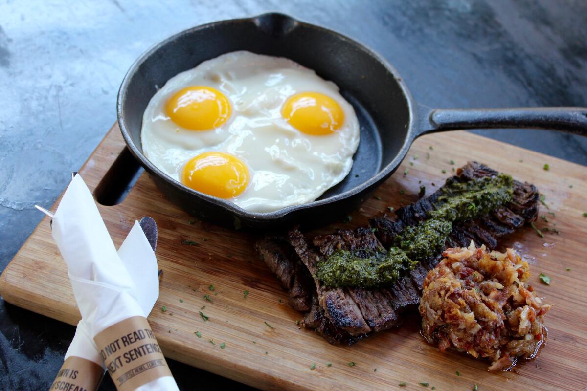 A steak, a breakfast pastry and a skillet with three sunny-side-up eggs sit atop a wooden cutting board. 