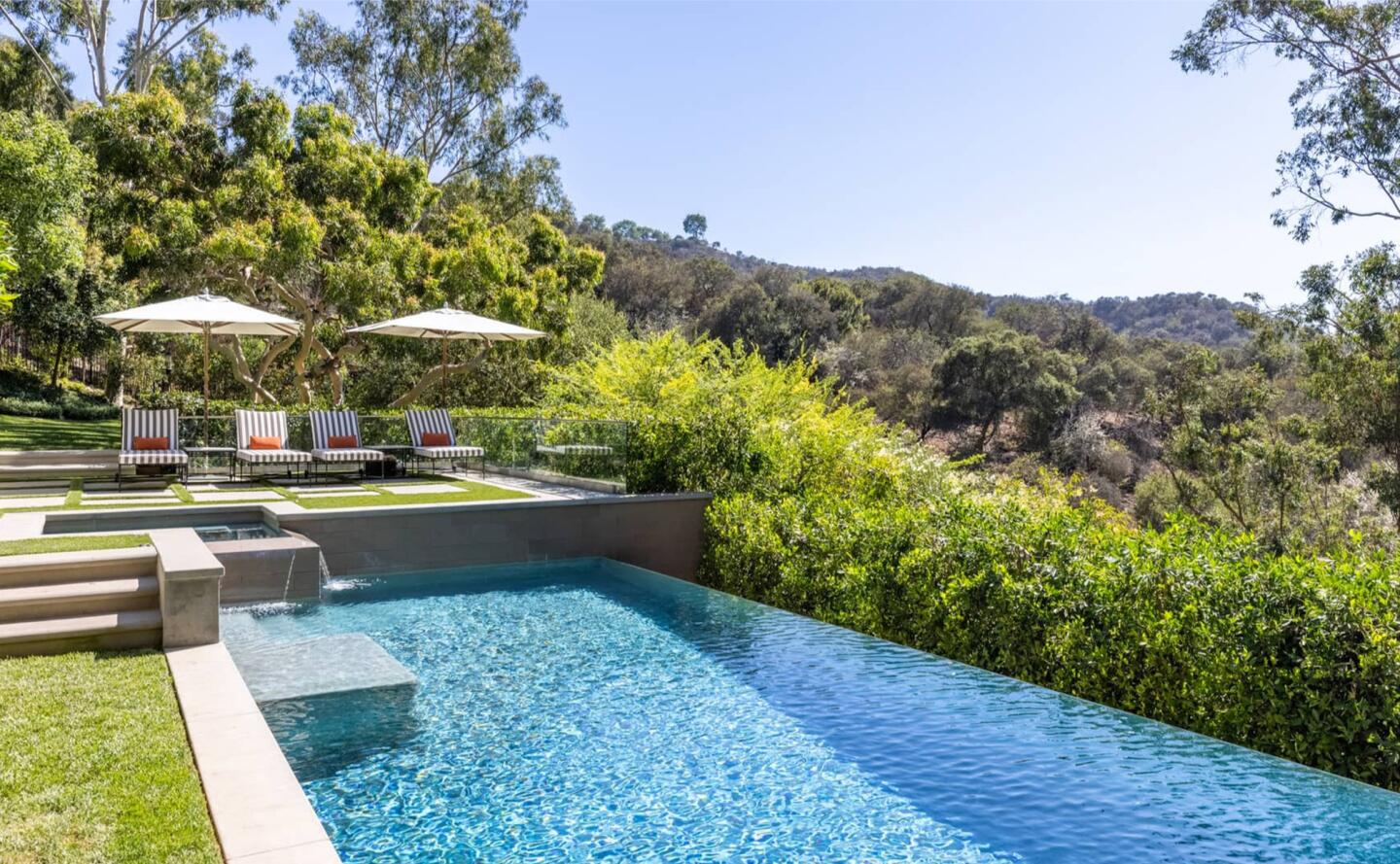 The pool has grass on one side and shrubs on the other side looking out over hills.