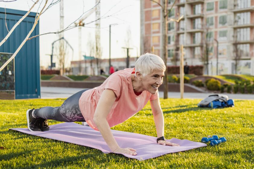 A smiling senior woman is doing pushups outside on a yoga mat.