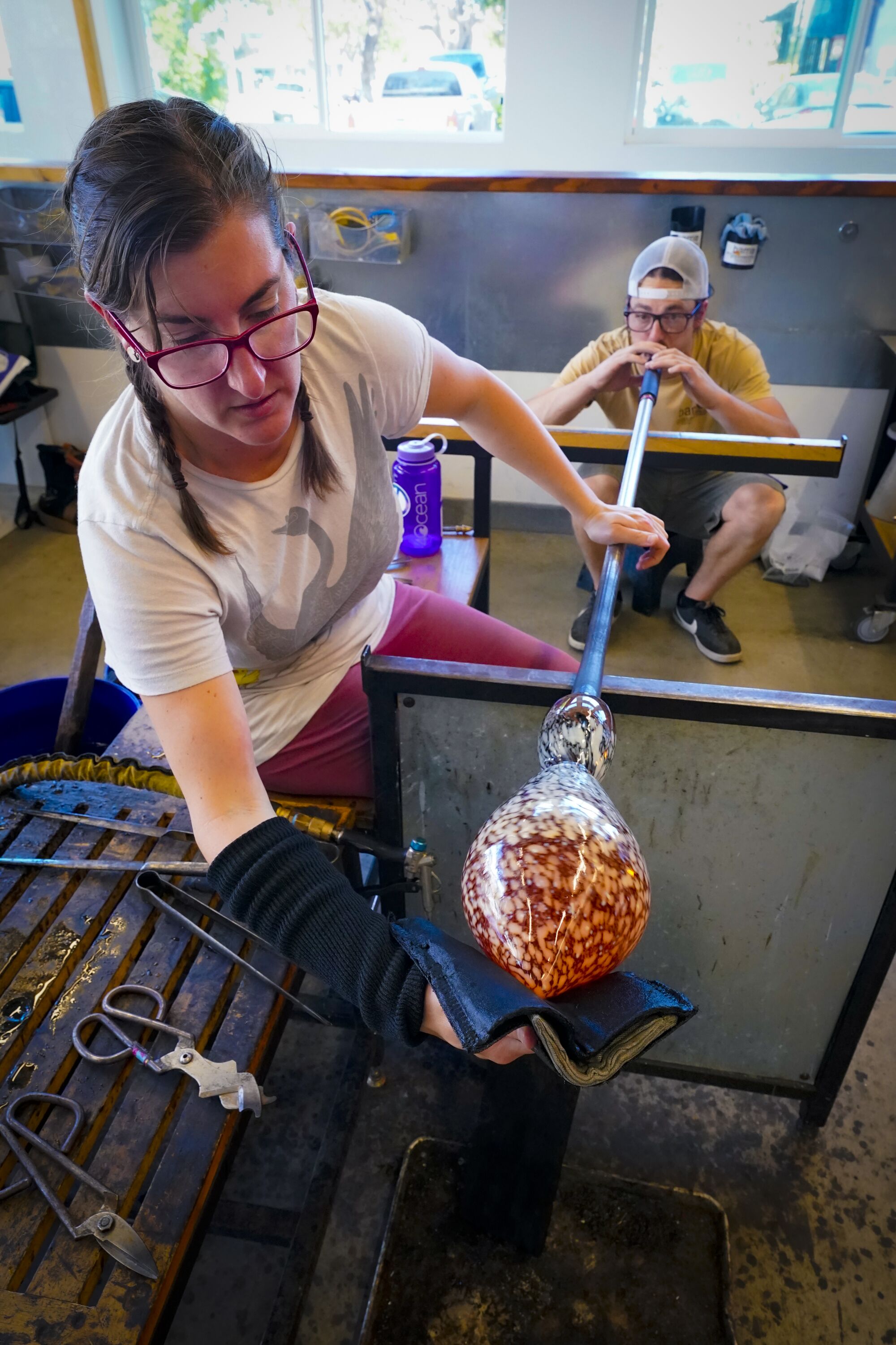 A glass artist works to shape a bowl, helped by an assistant blowing into the glass through a tube.