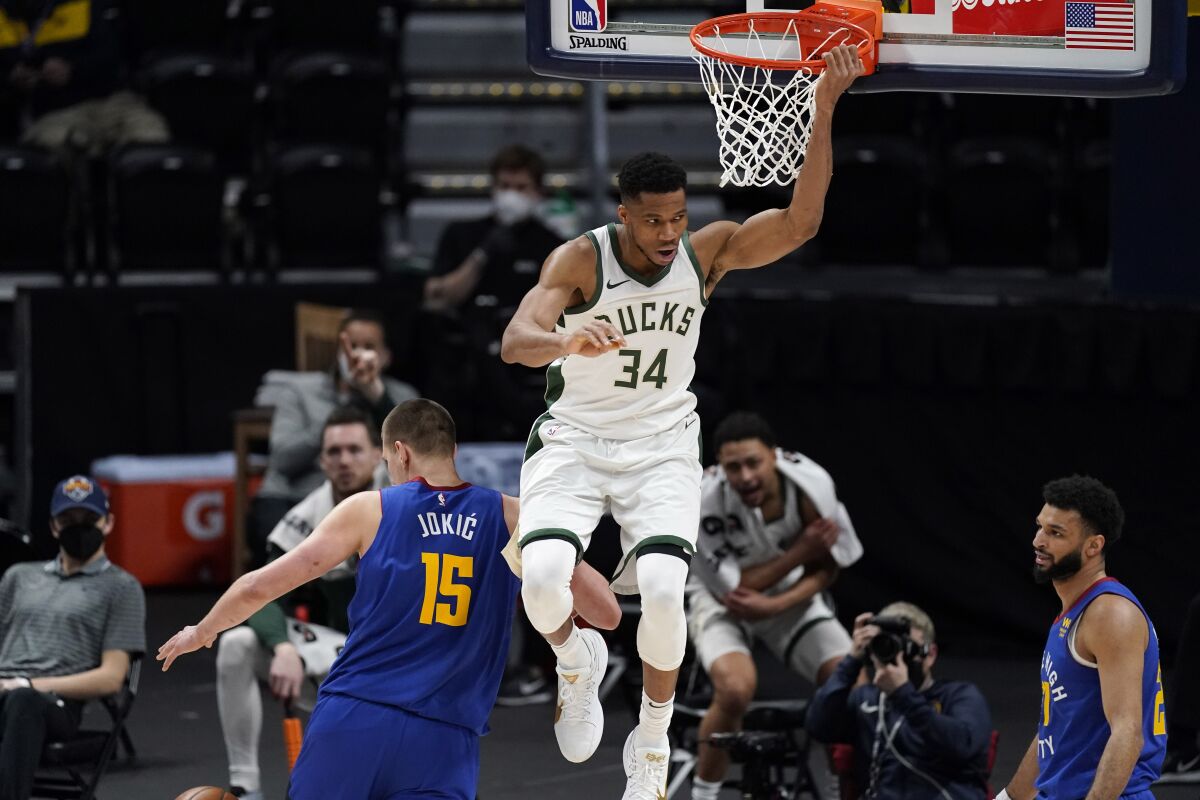 Milwaukee Bucks Giannis Antetokounmpo hangs on the rim after a dunk between Denver Nuggets Nikola Jokic and Jamal Murray.