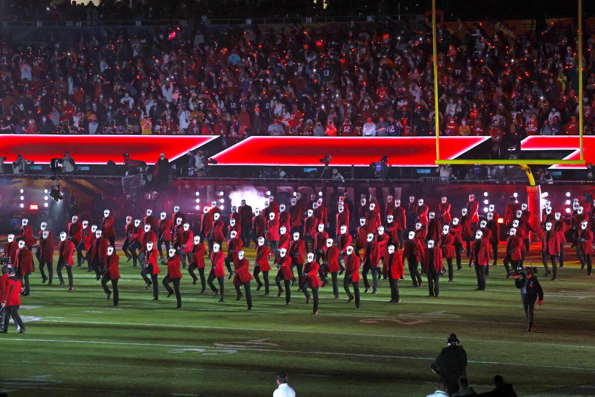 Performers in bright red jackets and white face masks march into a football field for a halftime show