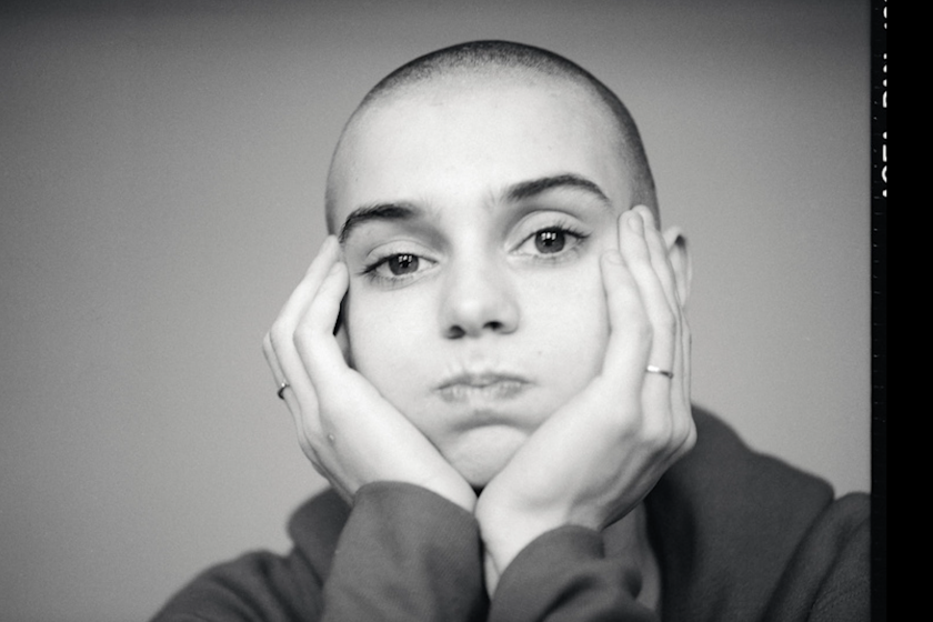 A black and white photograph of a young woman with a shaved head, resting her face in her hands