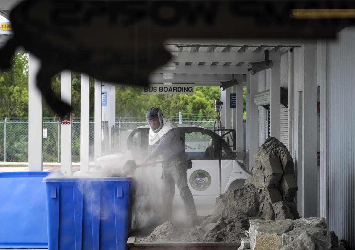 A worker shovels crushed basalt dust into a trash can while wearing a protective hood.