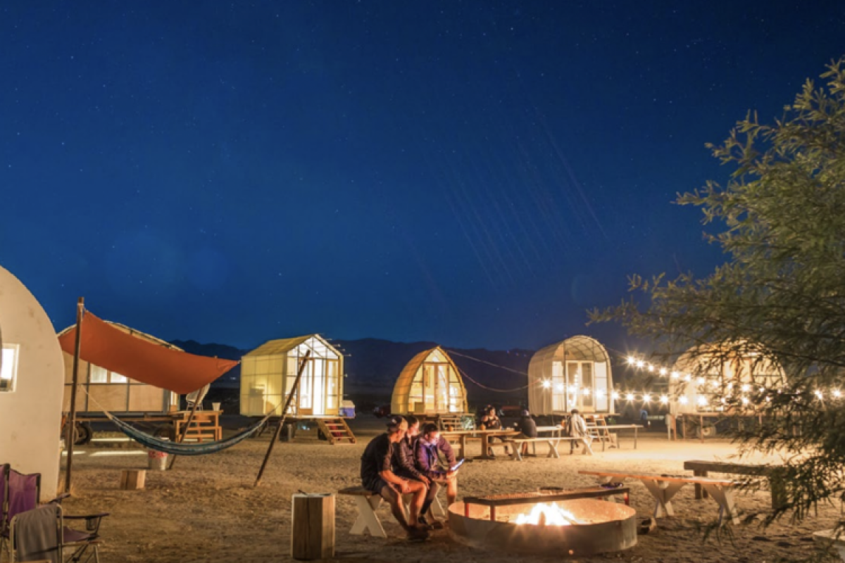 Outdoor glamping huts lit up under a night sky, with people in the foreground next to a fire pit