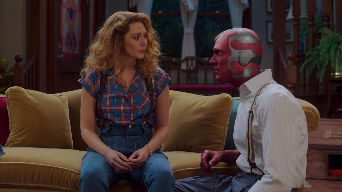 A woman with red hair sitting on a couch and staring at a humanoid robot kneeling at her side.