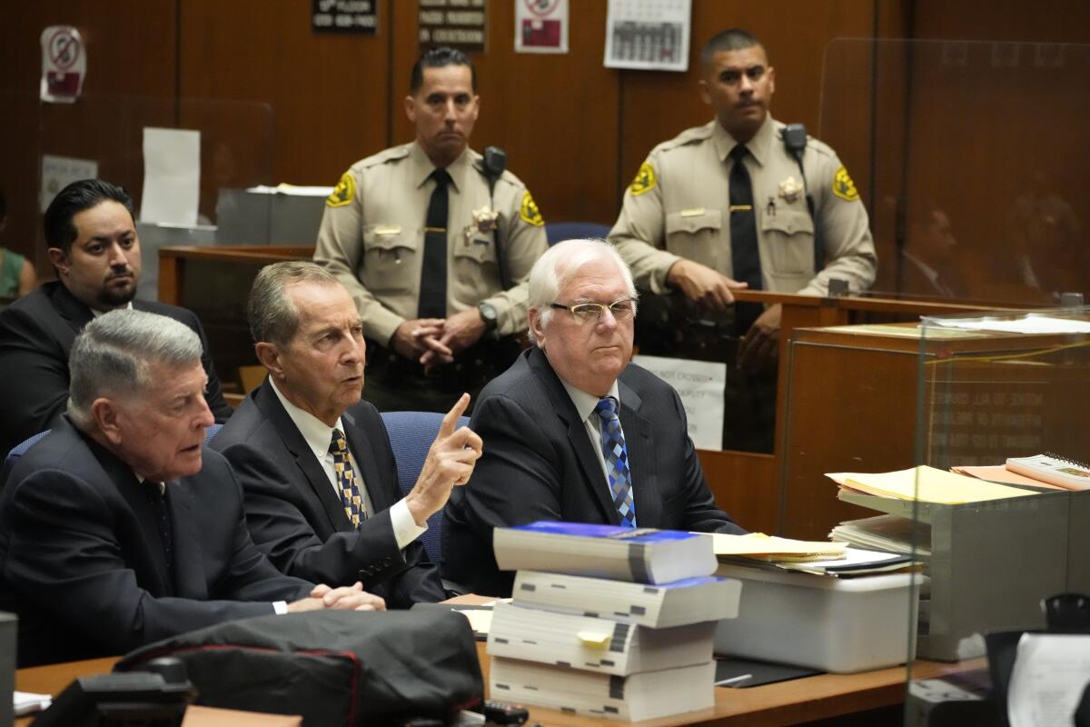 Three men sit a table with two men in uniform behind them in a courtroom.