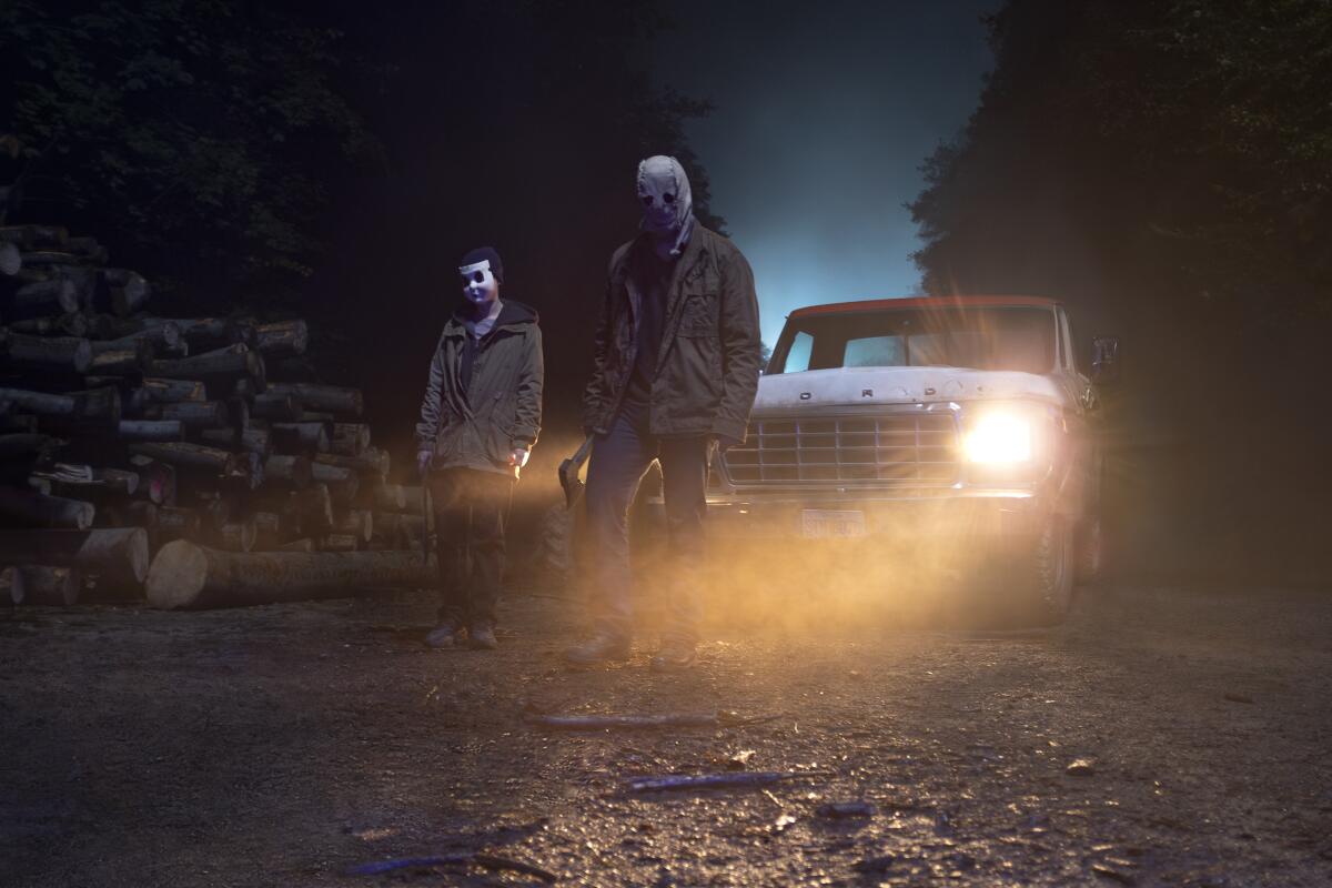 Two masked characters stand in front of a truck with its headlights on in "The Strangers - Chapter 1."
