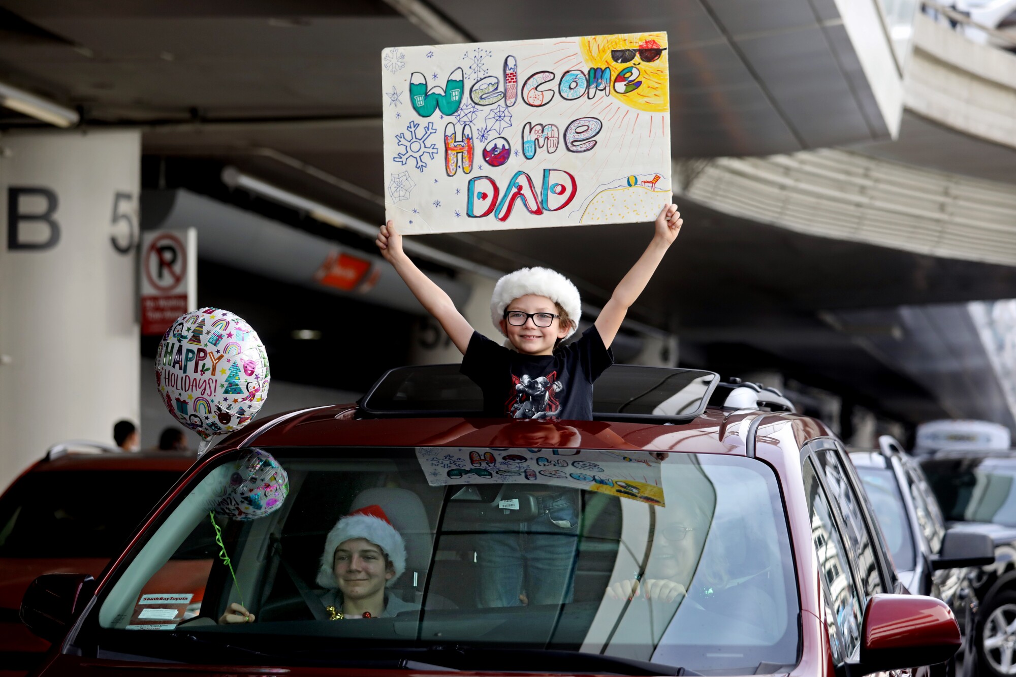A boy holds up a sign saying Welcome Home Dad inside a car with a balloon out the window