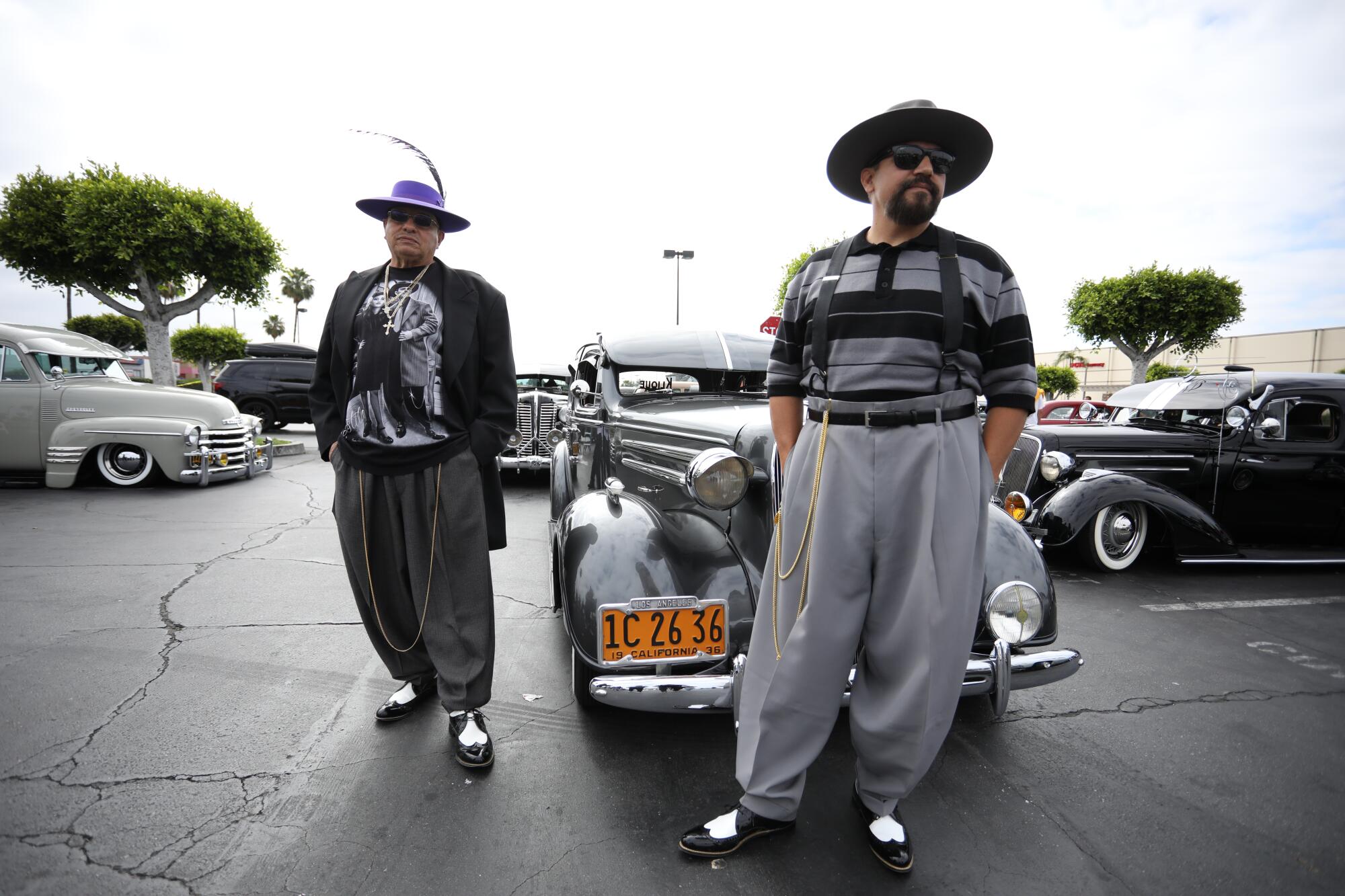 Two men in pants pegged at the ankle, with long chains, hats and shining saddle shoes, stand near classic cars.