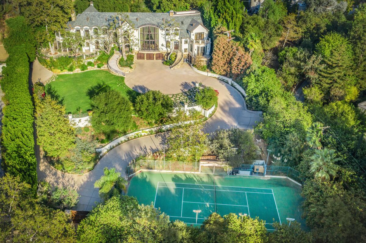 An aerial view of a two story mansion surrounded by trees with a nearby tennis court.