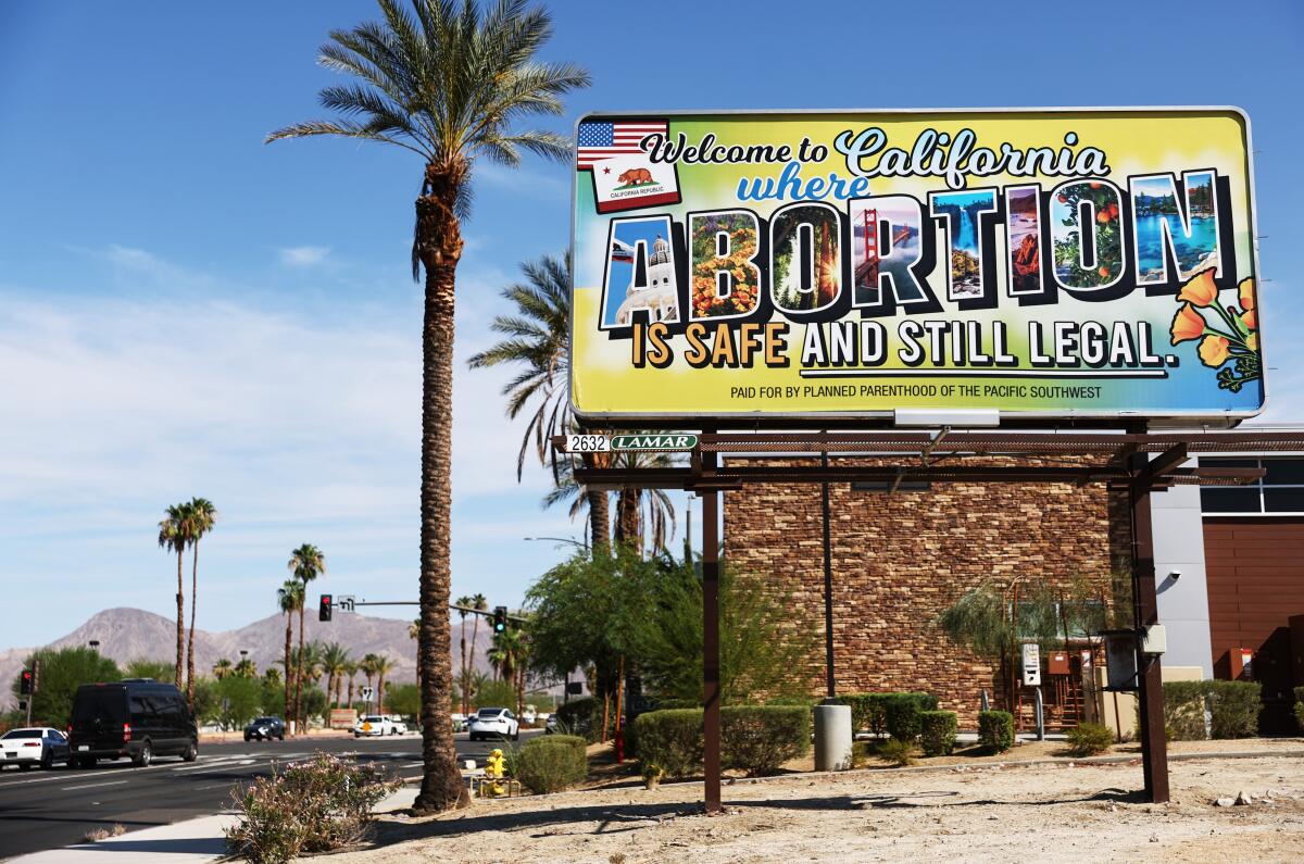 A billboard stands next to a palm tree, alongside a city street with palm trees and mountains in the distance.