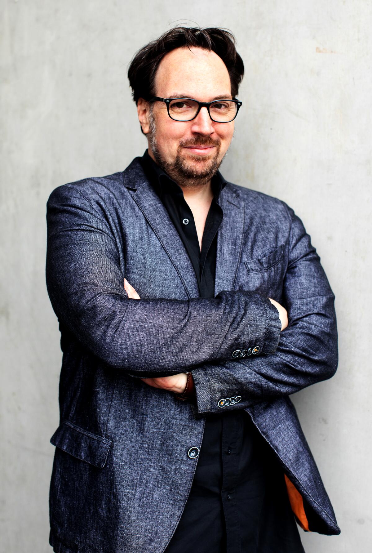 An author stands with his arms crossed, wearing glasses, a shiny gray jacket and a black shirt open at the collar.