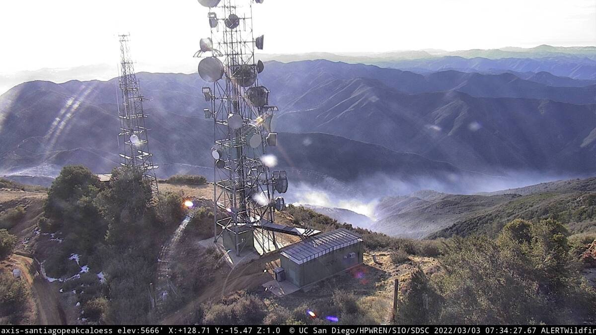 View from a wildfire camera shows smoke in a canyon with communications towers in the foreground.