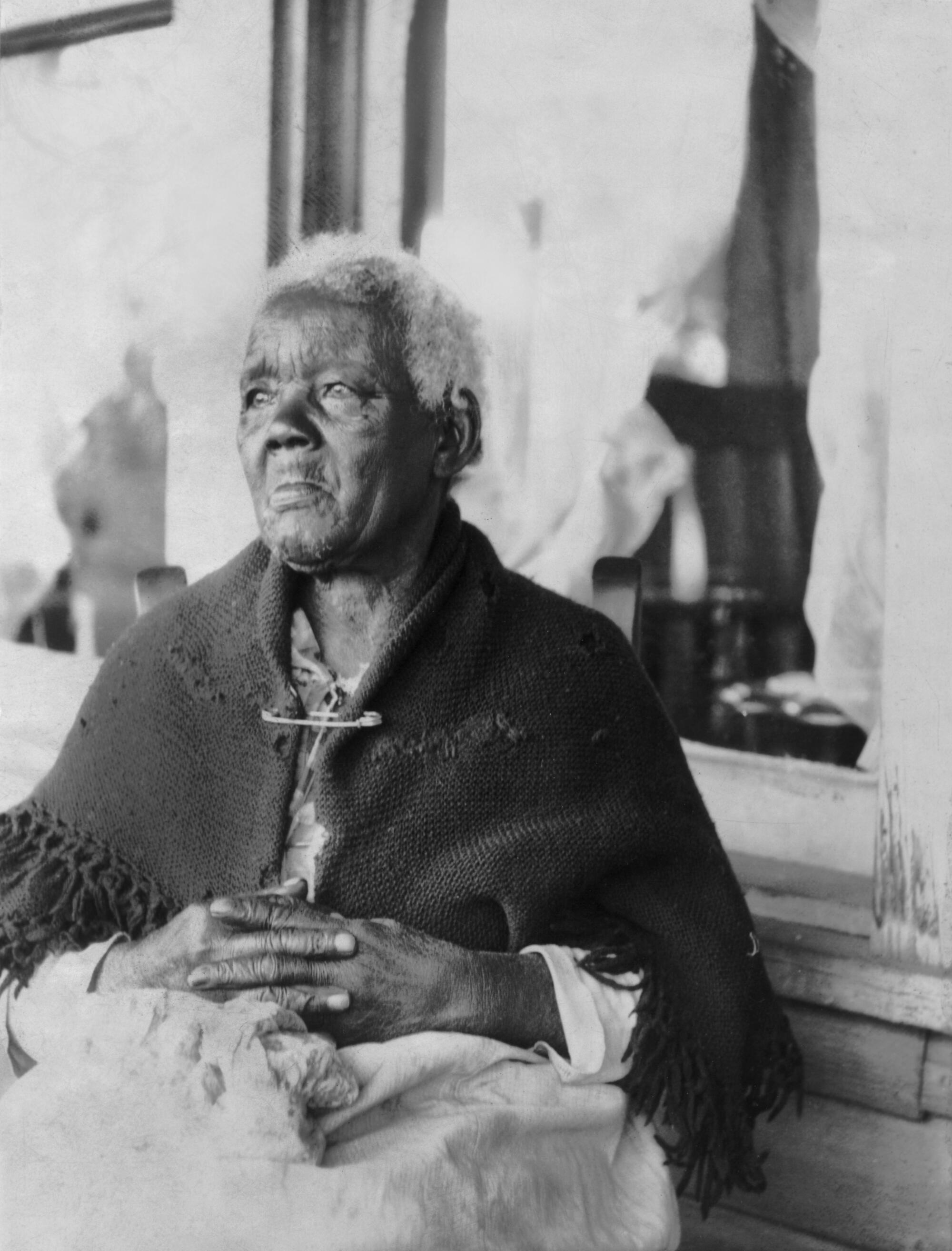 A woman sits in front of a curtained window, wearing a shawl, her fingers interlaced in her lap.