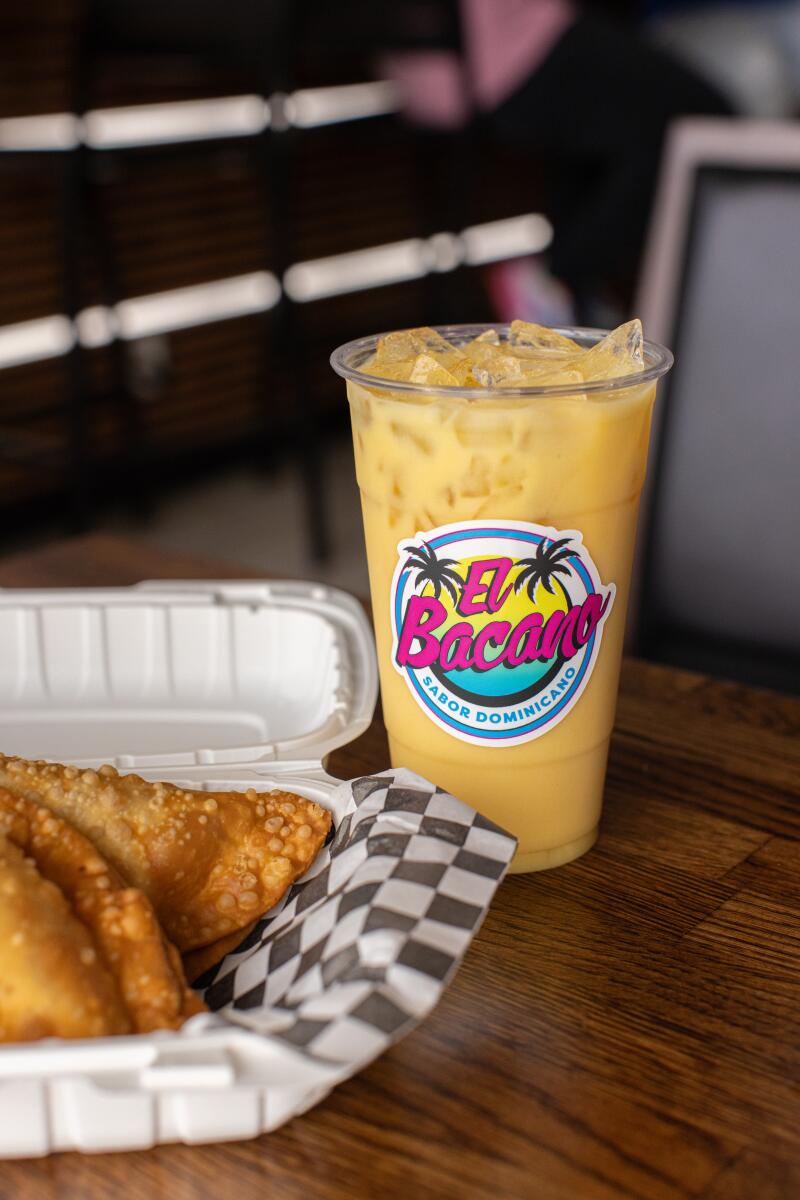 A plastic cup with the El Bacano logo is filled with ice and a golden liquid, next to a container of empanadas. 
