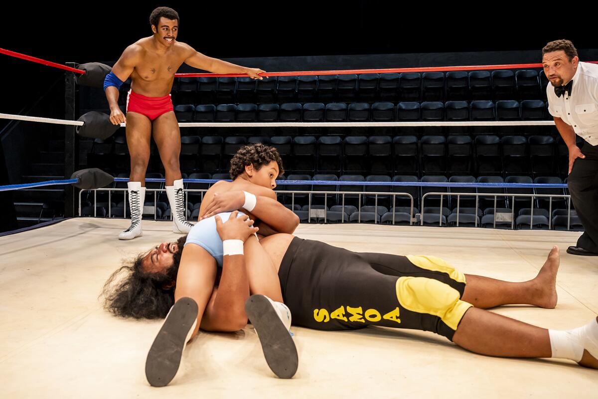 In a pro wrestling ring, a wrestler pretends to be pinned by a young boy as another wrestler looks on.