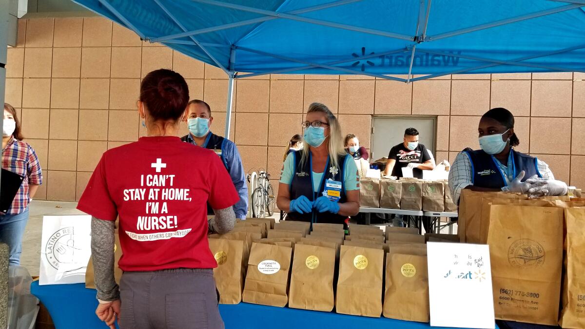 Volunteers stand at a table lined with brown bags containing food for distribution.