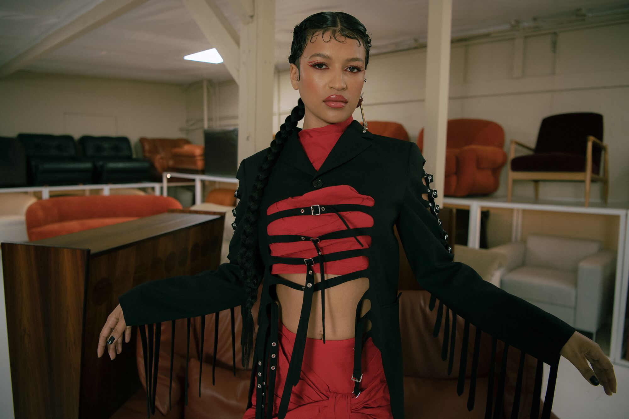 A model wearing a red and black outfit looks at the camera, chairs and couches in the background.
