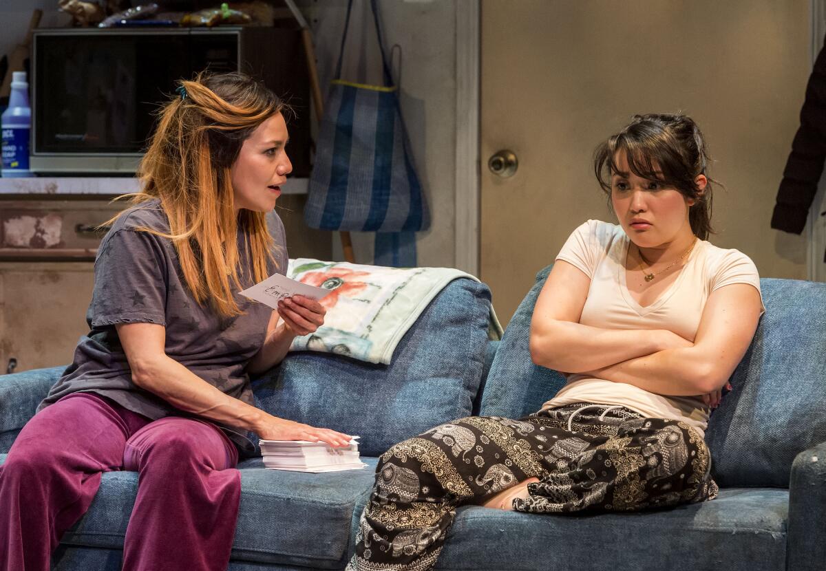 A woman sitting on a couch speaks to another woman on the couch, who crosses her arms and looks angry 