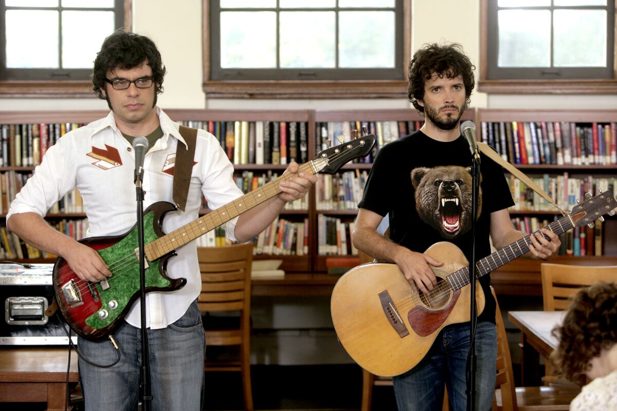 Two men playing guitars and standing at microphones in front of a bookshelf.