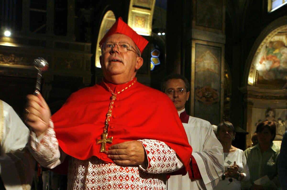 A man with glasses, wearing a red hat and a cross over a red half-cape and religious robe, holds an aspergillum 