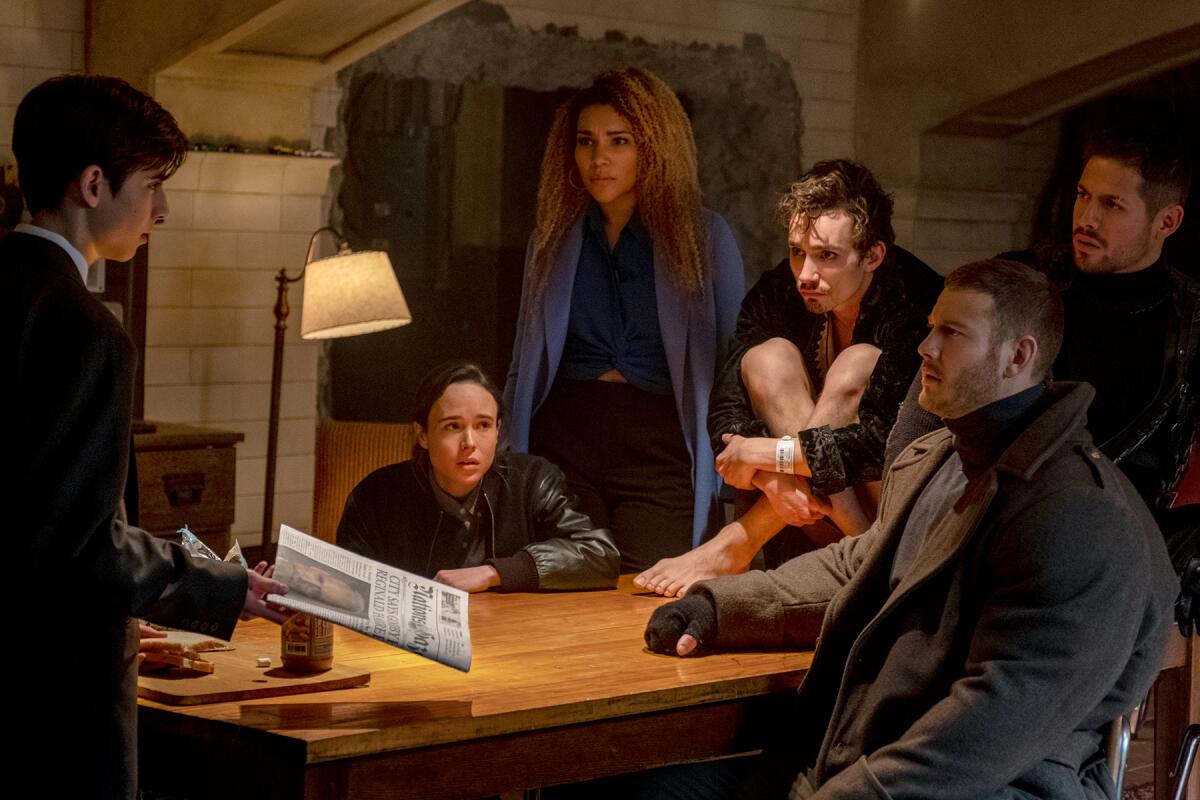 A boy holds a newspaper and five people at a table look at him