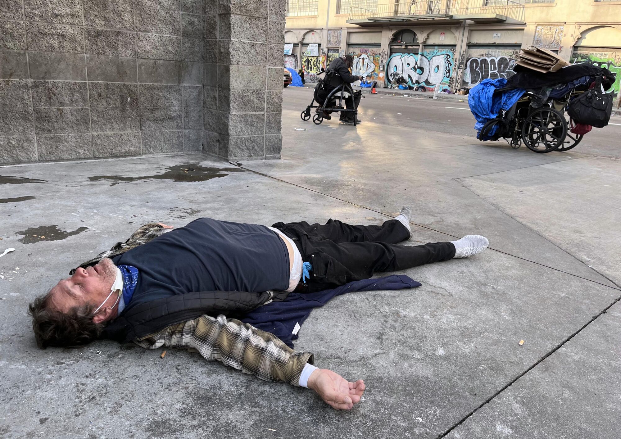 A homeless man is passed out on a sidewalk near a wheelchair.