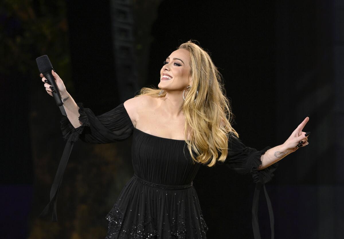 Adele, in a black dress, smiles and holds a microphone in one of her outstretched hands.