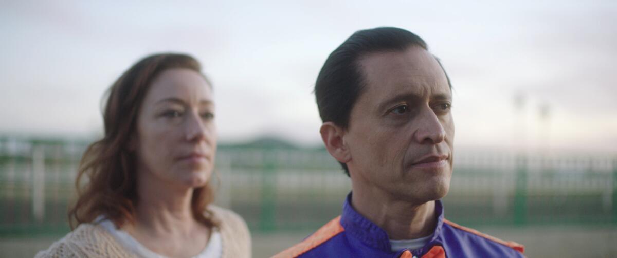 Closeup of the faces of a woman and man outdoors near a racetrack.