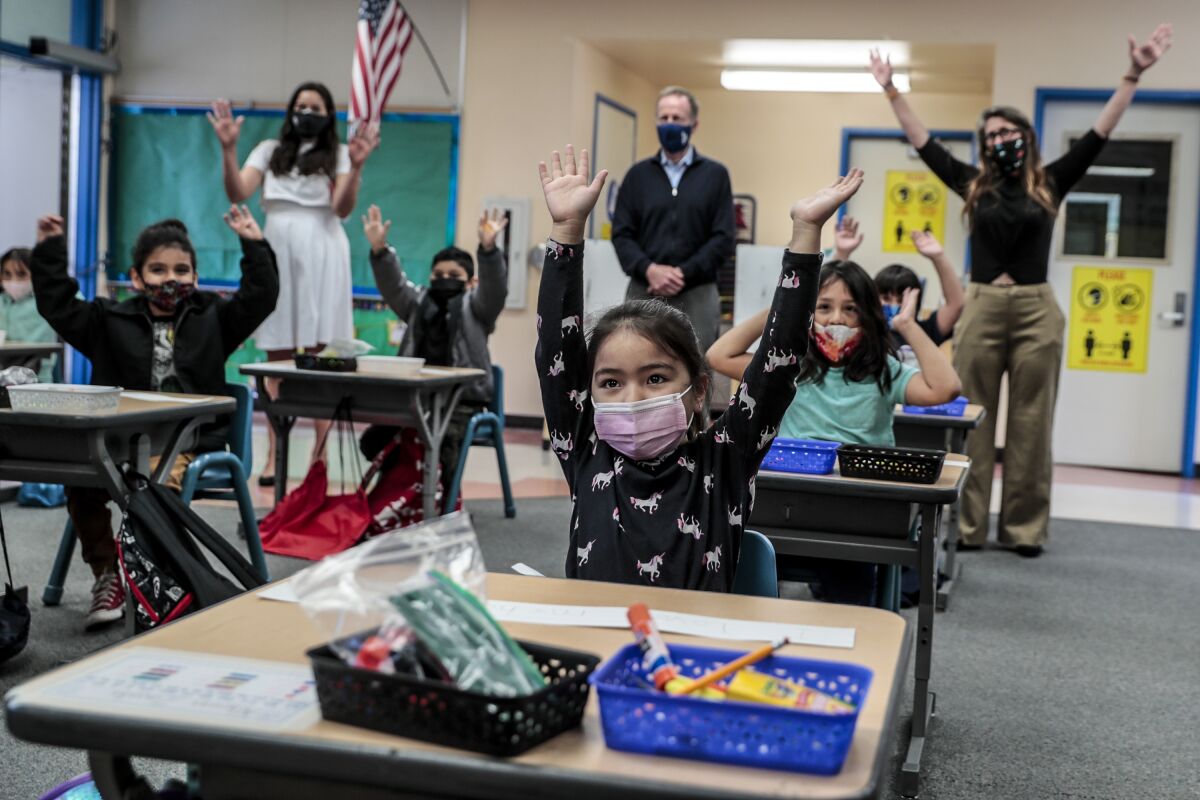 Kindergarten children in masks sitting at desks raise their hands for a photo