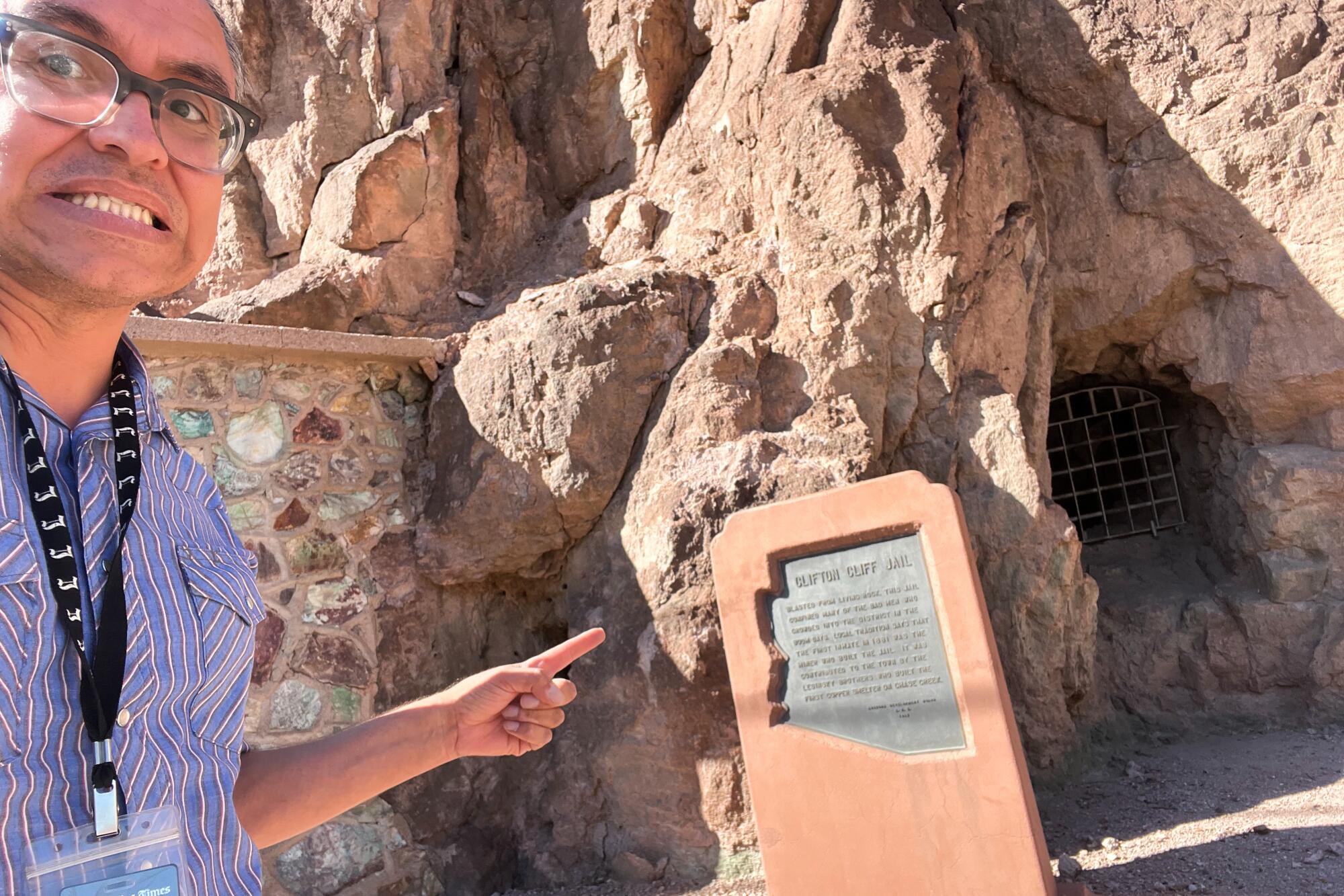 A man with glasses points to a plaque outside a rocky facade with a small opening covered by a metal grille