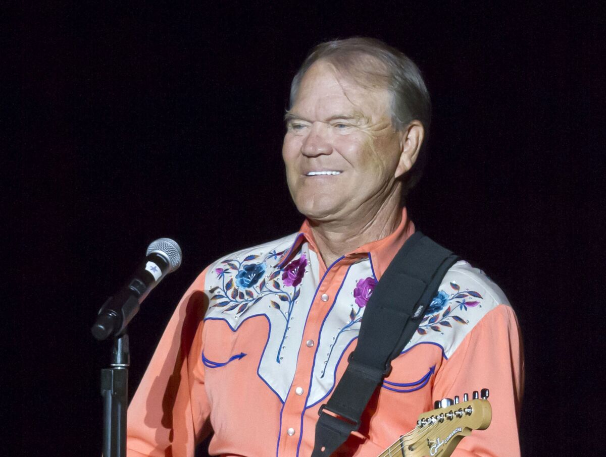 A man in an embroidered cowboy shirt smiles and plays guitar