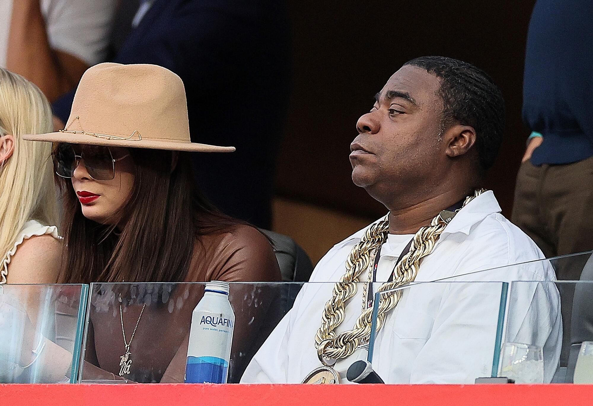 A woman in a hat and sunglasses sits next to a man in a white shirt and large gold chain at a football game.