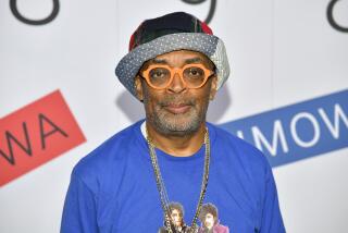 Spike Lee in thick orange glasses, a patterned bucket hat, bright blue shirt and a long gold chain looking straight ahead