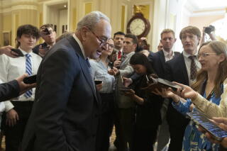 A man with gray hair and glasses, in a dark suit, speaks to people holding phones toward him