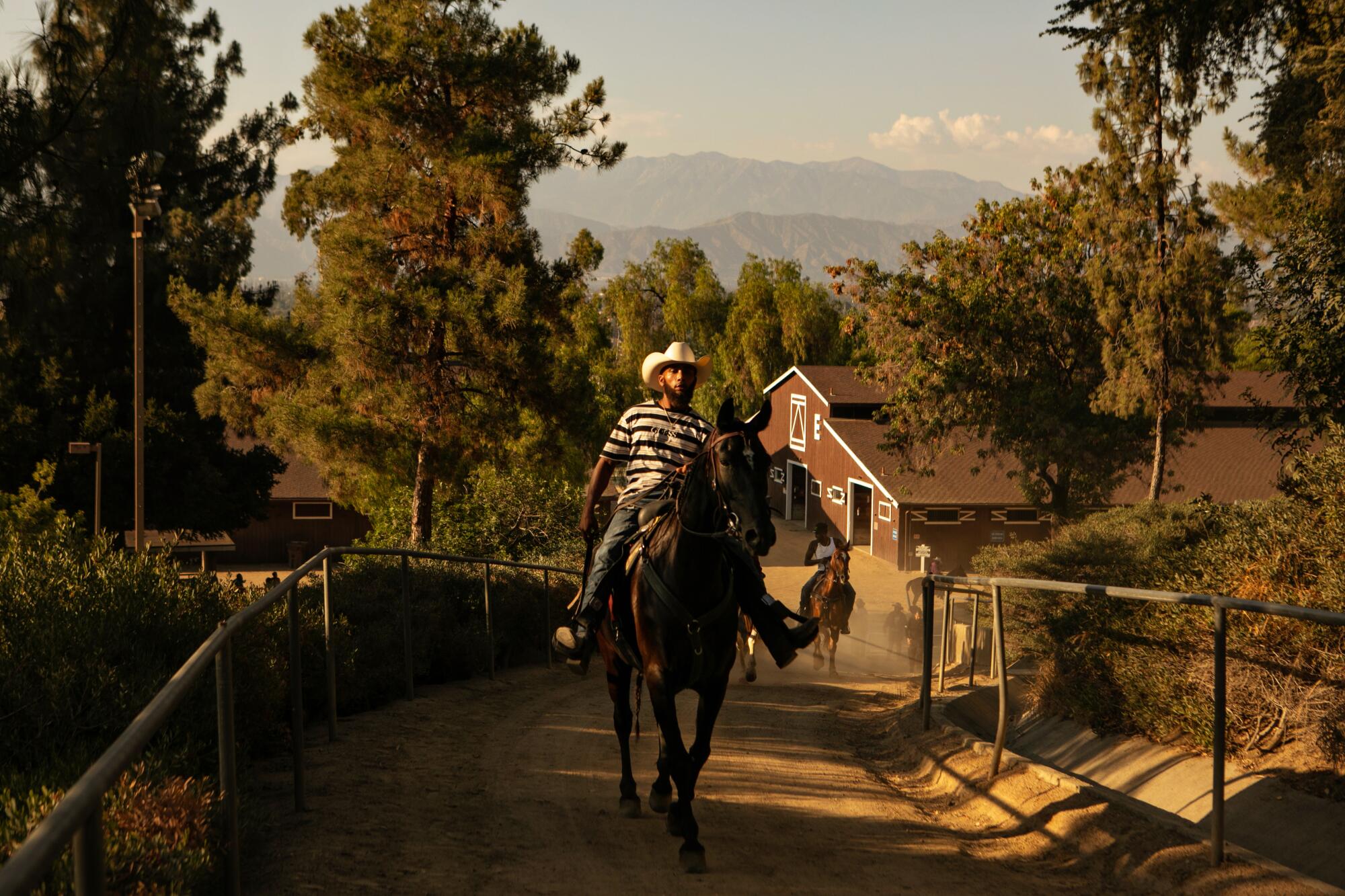 Black men riding horses on a dirt road, with a stable, evergreens and mountains in the background